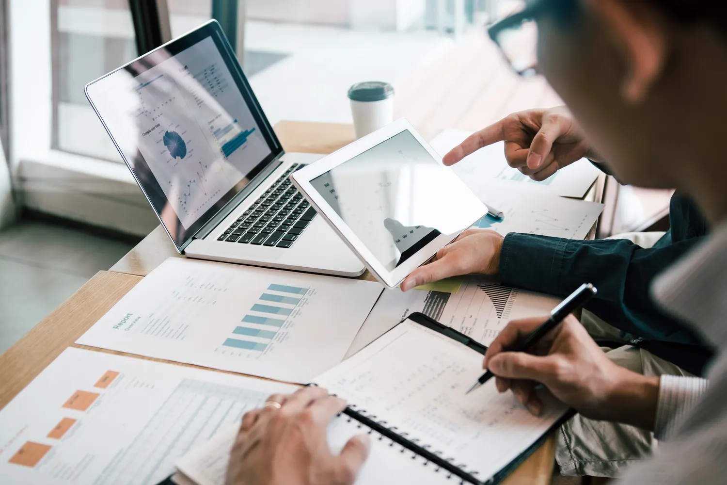 Burow Finance - Consulting GmbH - Wirtschaftsexperten arbeiten gemeinsam an einem Finanzanalyseprojekt und nutzen dafür Laptop, Tablet und gedruckte Berichte.